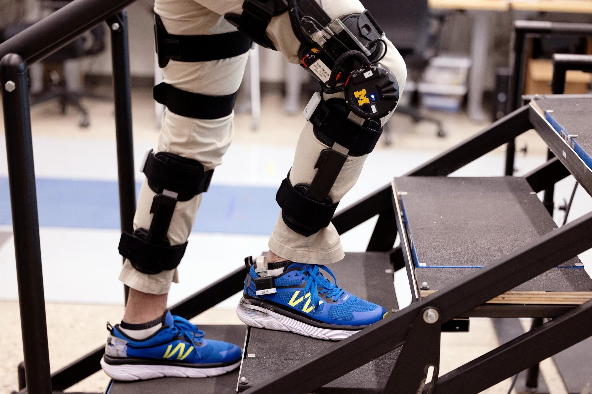 A person wearing a powered knee exoskeleton module climbing a laboratory staircase.