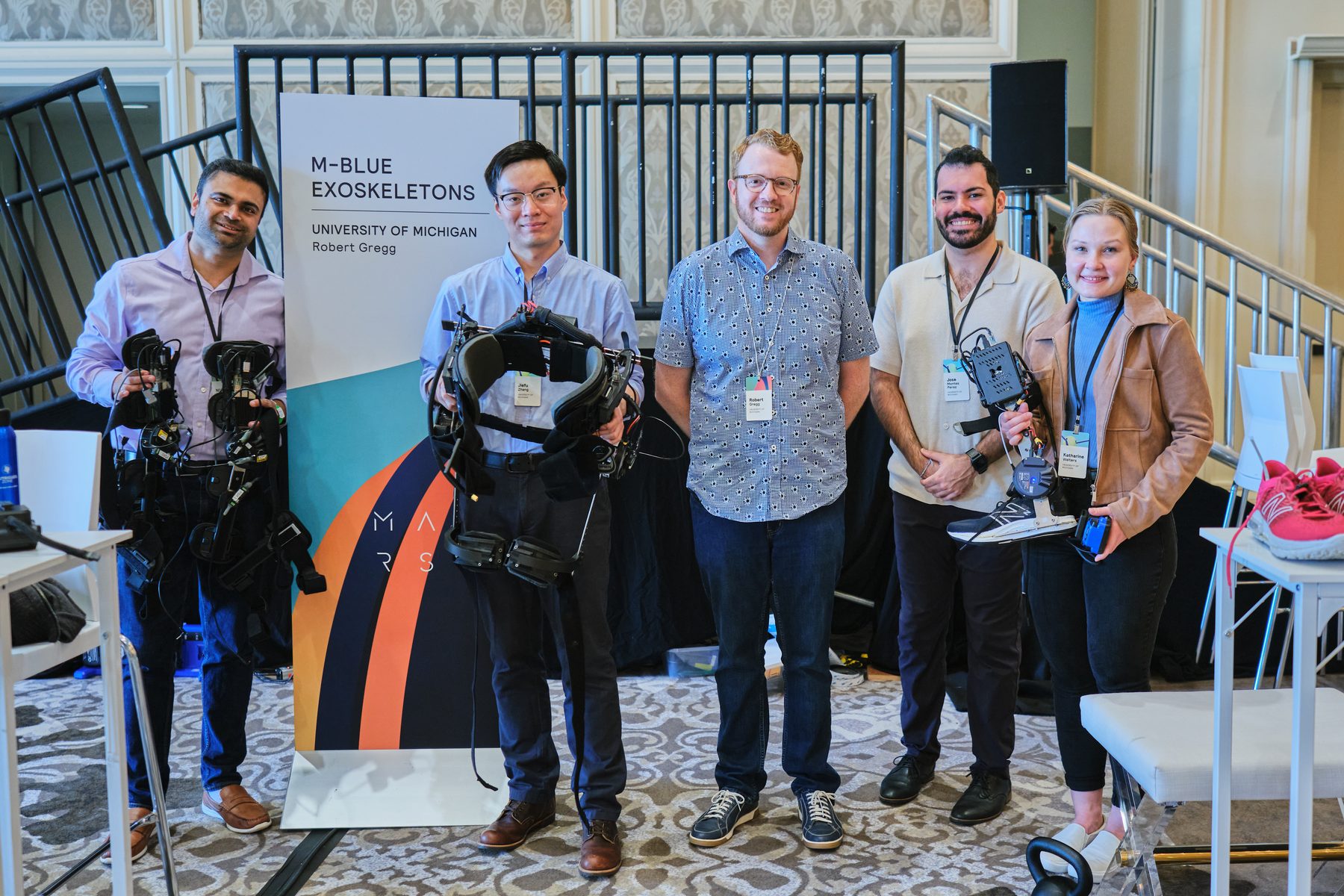 Five lab members standing with M-BLUE exoskeleton hardware at a conference exhibit.